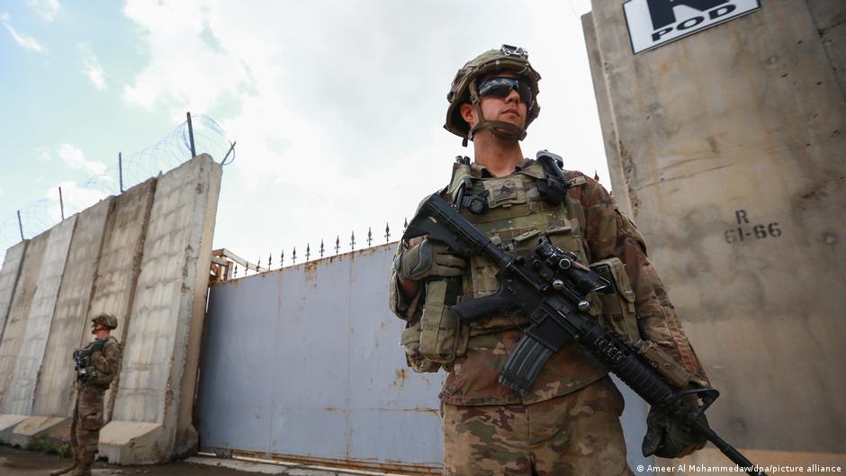 A soldier in camouflage and protective gear holds a rifle beside a concrete gate, with another soldier in the background.