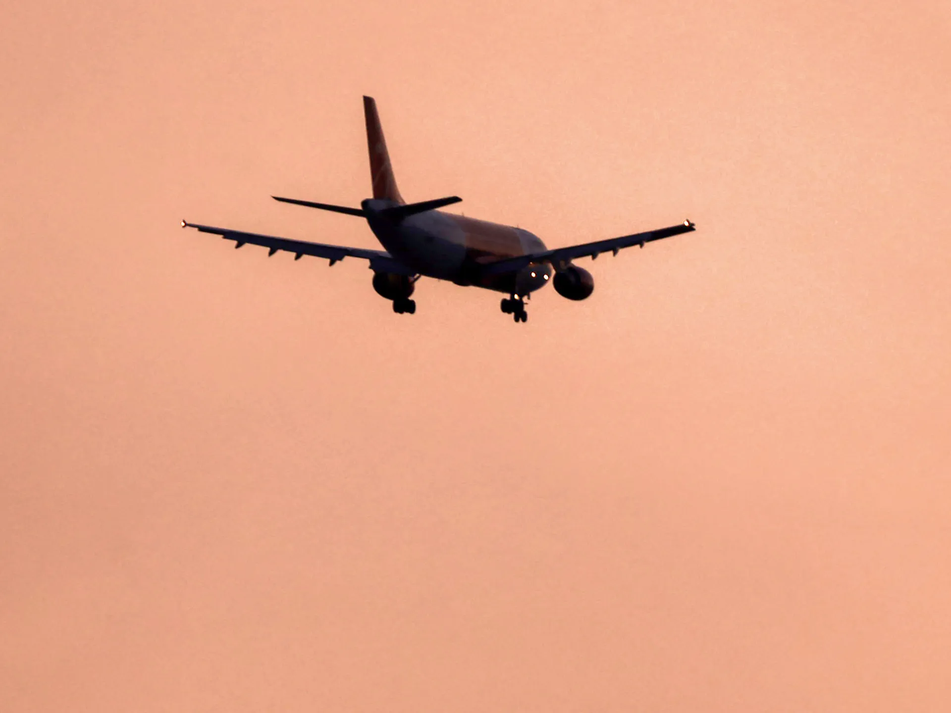 A passenger jet descends with landing gear lowered against a peach-colored sunset sky.