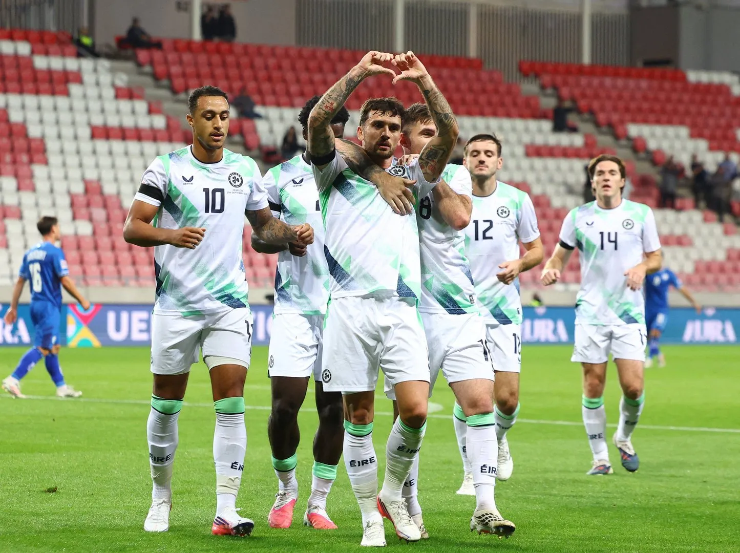 Irish players celebrate a goal on the pitch, one making a heart shape with his hands.