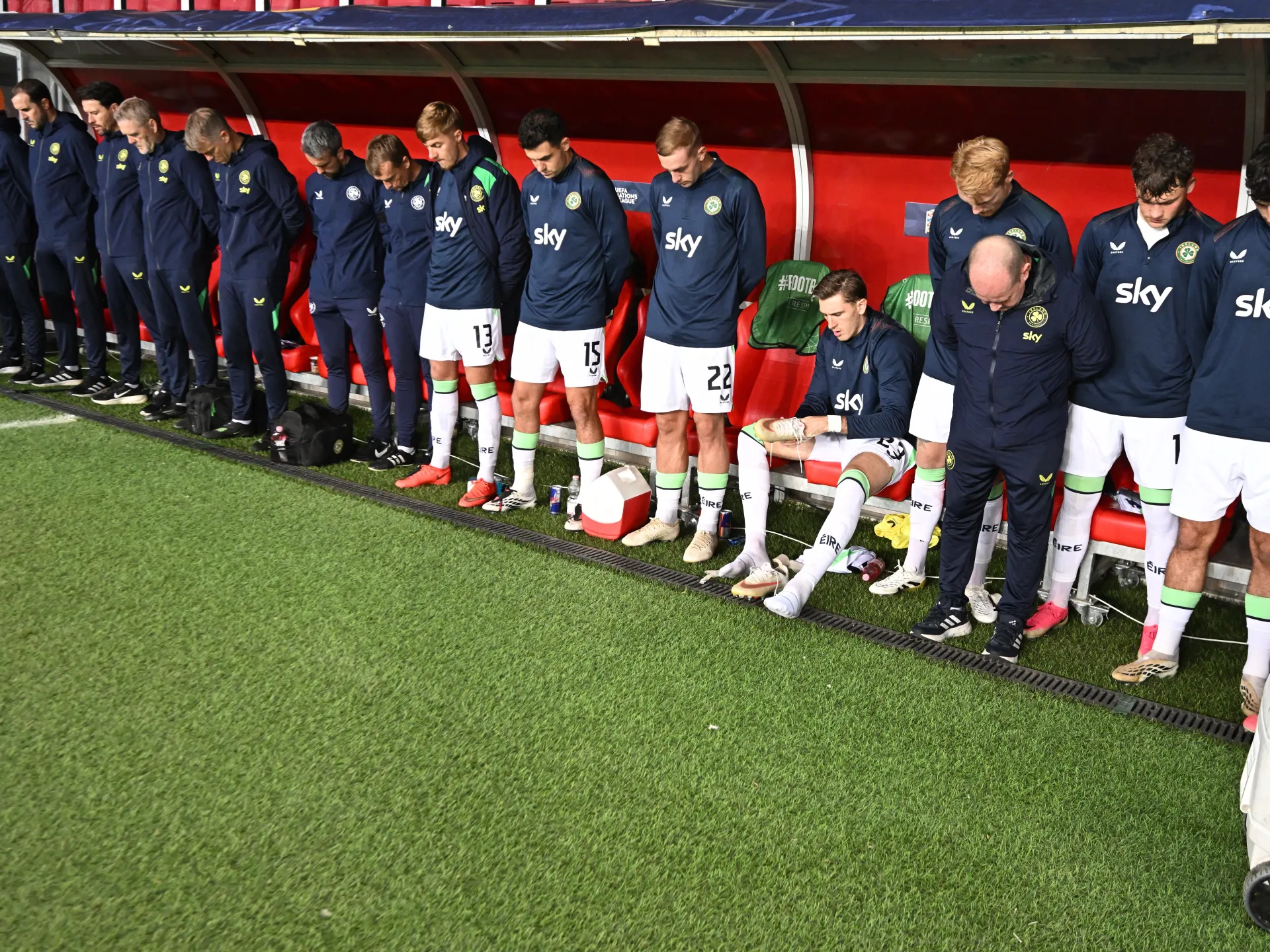 Irish football players and staff stand solemnly beside the dugout, with one player seated and tending to his boot.