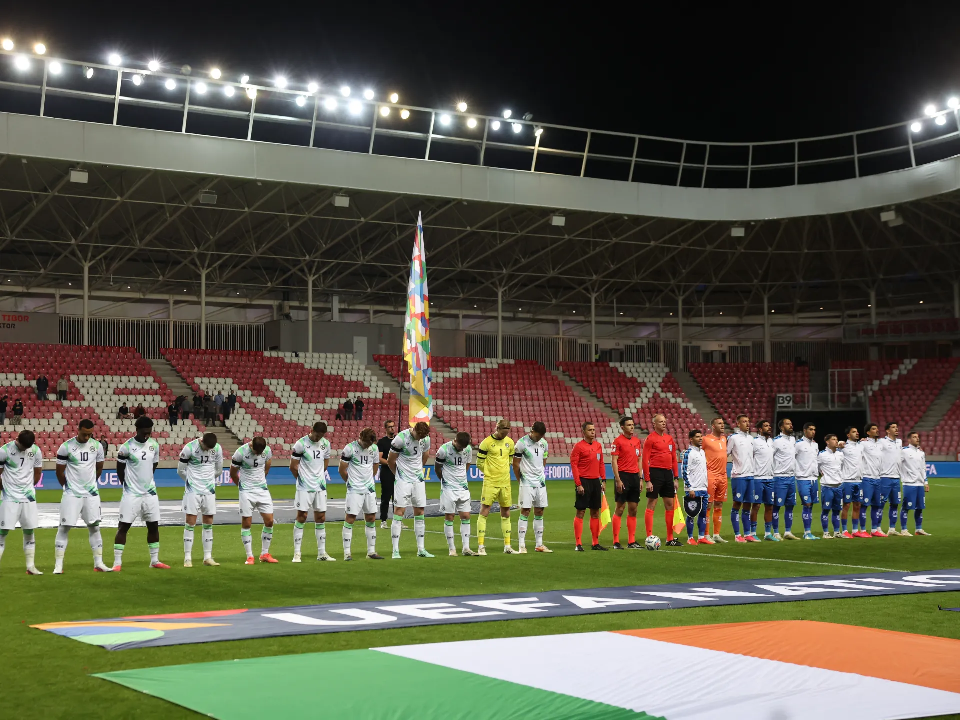 Football teams stand in line on a floodlit pitch, with flags laid across the grass.