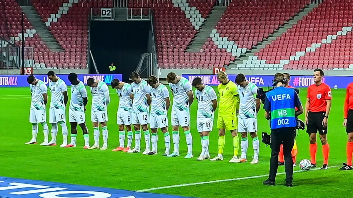 Football players in white kits stand with heads lowered beside a goalkeeper during a pre-match ceremony.