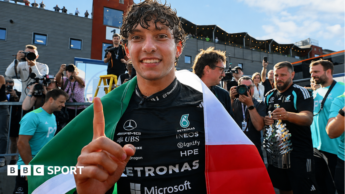 Smiling racing driver gestures beneath an Italian flag amid photographers and Formula One team staff.