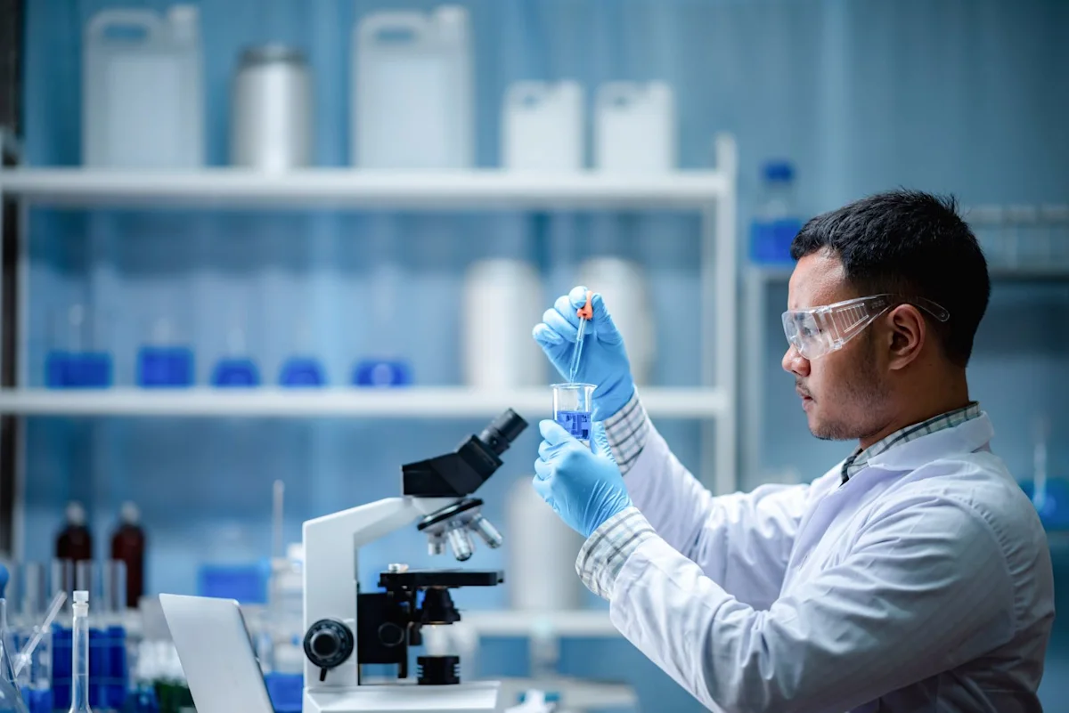 Scientist in protective eyewear and gloves uses a pipette beside a microscope in a laboratory.