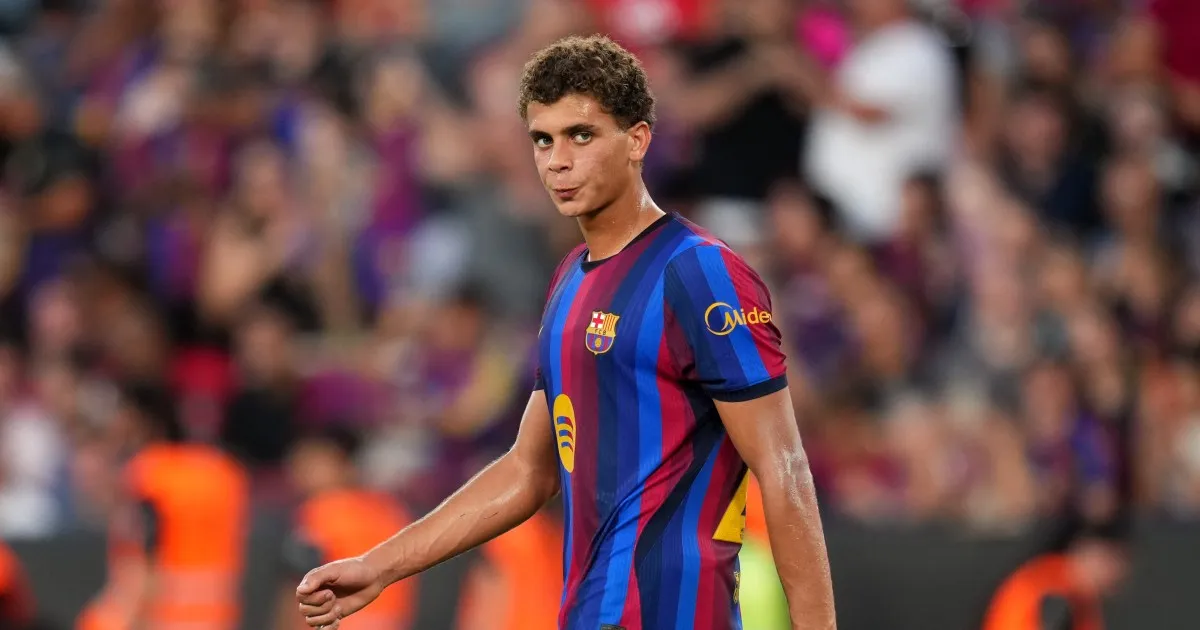 Footballer in Barcelona colors walks across the pitch with a blurred crowd behind him.