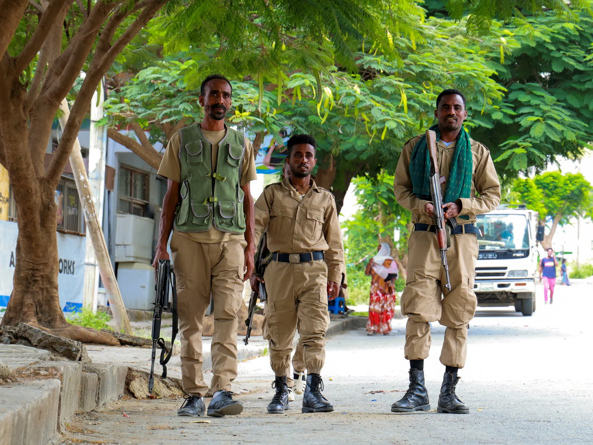 Three armed men in tan uniforms walk beneath leafy trees along a street, with a truck behind them.