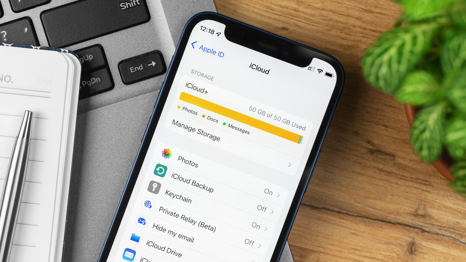 iPhone displaying iCloud storage settings beside a laptop, notebook, pen, and green plant