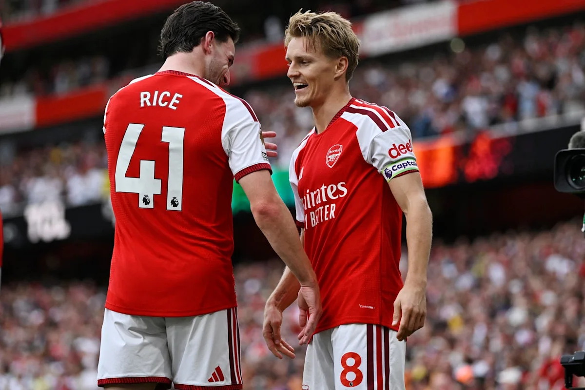 Two Arsenal footballers in red kits stand together during a match at a crowded stadium.
