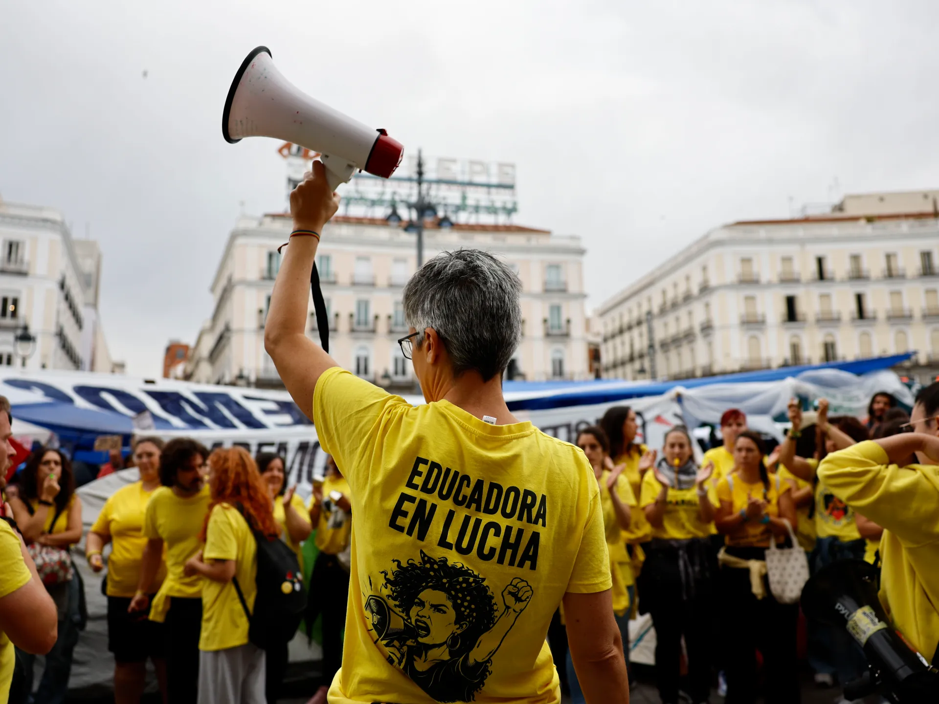 A protester in a yellow shirt raises a megaphone above a crowd in a city square.