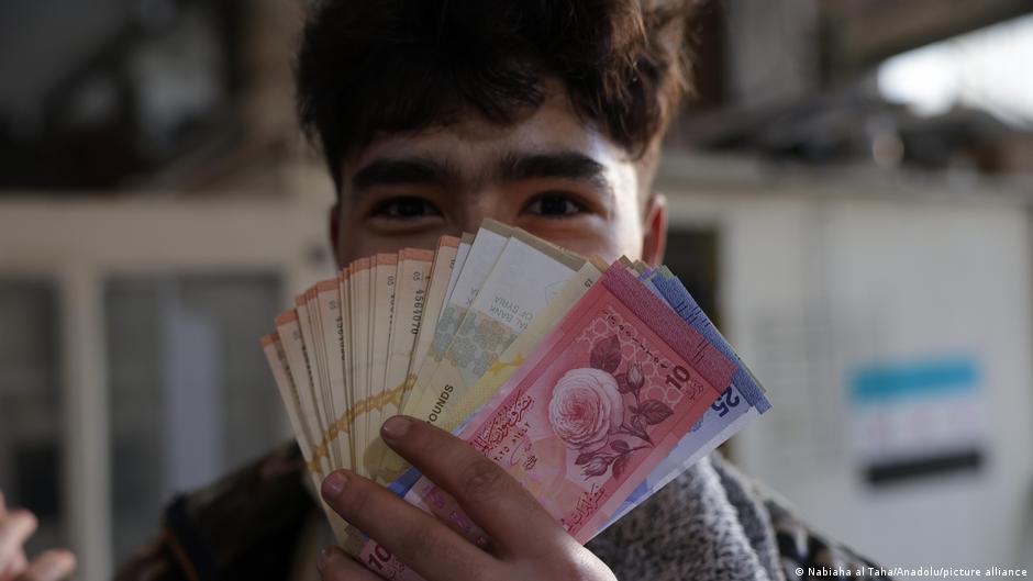 A young man holds several banknotes fanned across his face, with only his eyes visible.