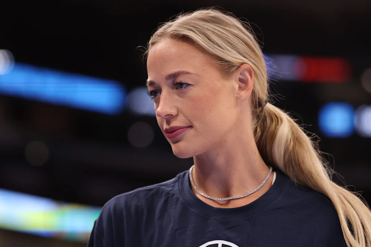 Blonde woman with ponytail wearing a dark shirt and necklace at a sports venue