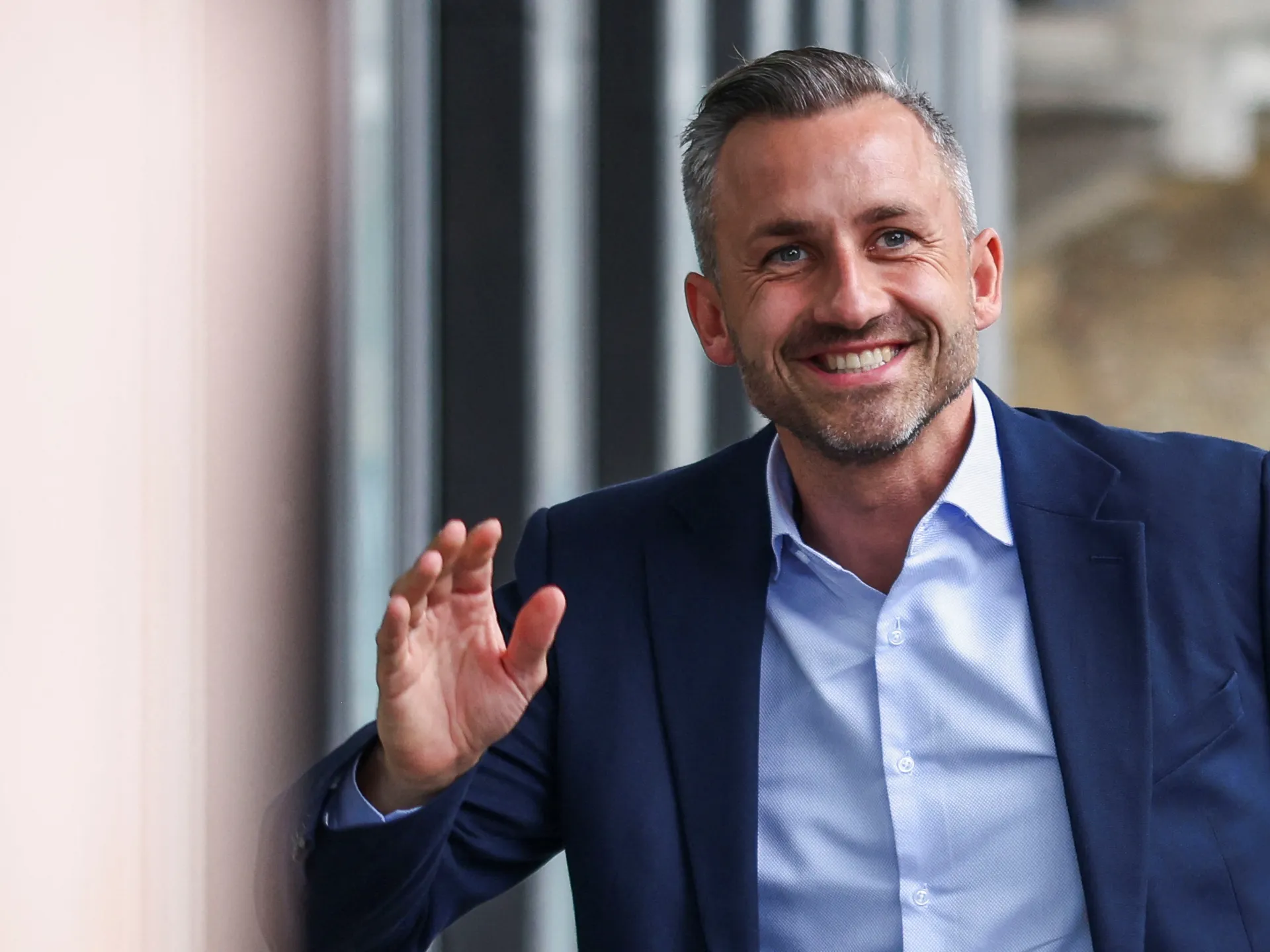Smiling man in a navy suit gestures while speaking indoors