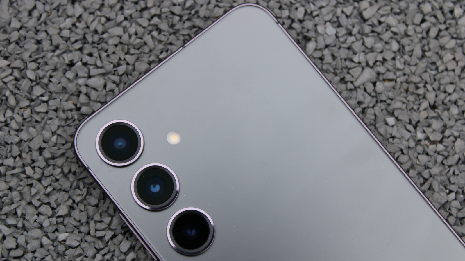 Close-up of a silver Samsung smartphone’s triple rear cameras resting on gray gravel.