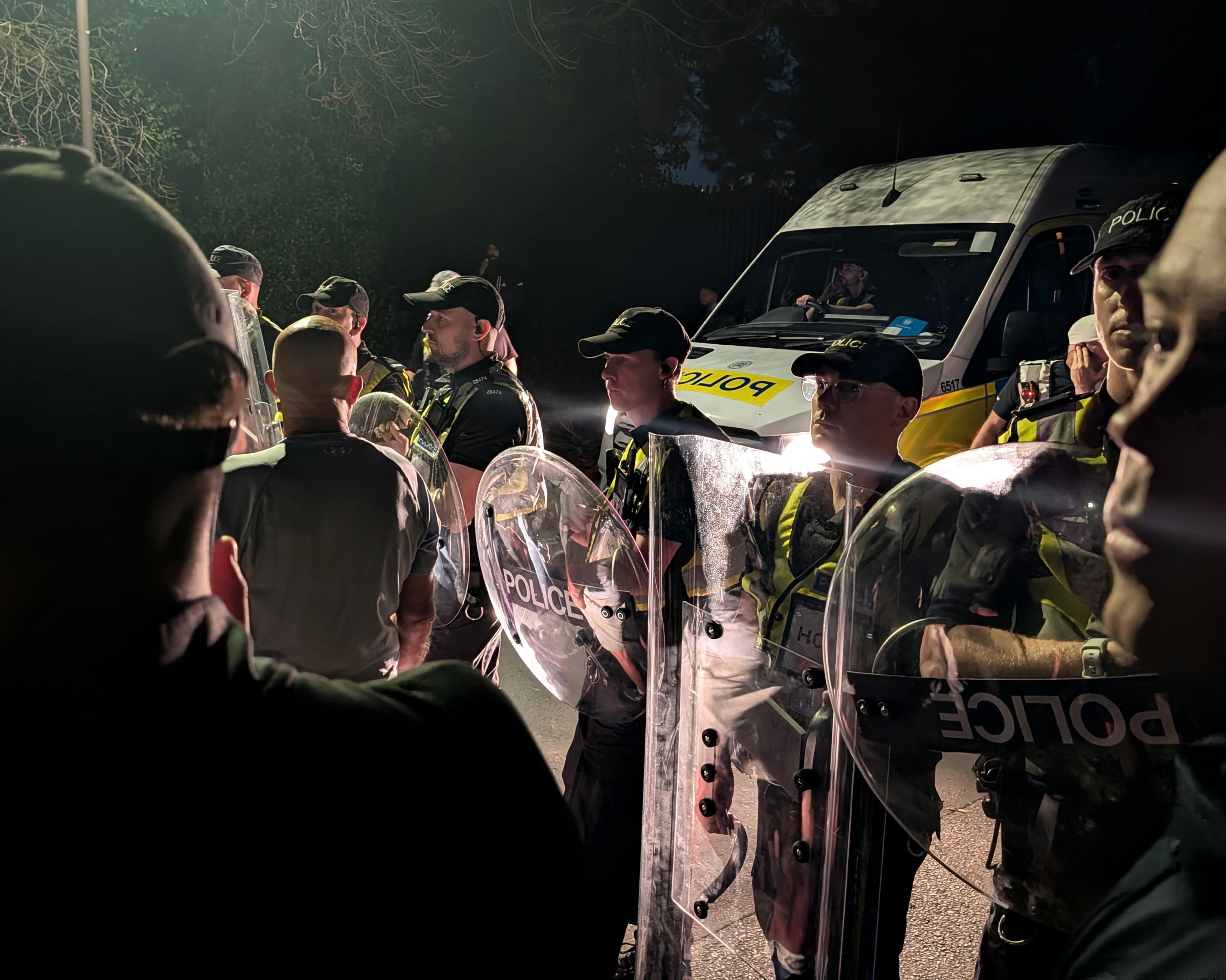 Police officers in riot gear stand beside a police van at night amid a crowd.