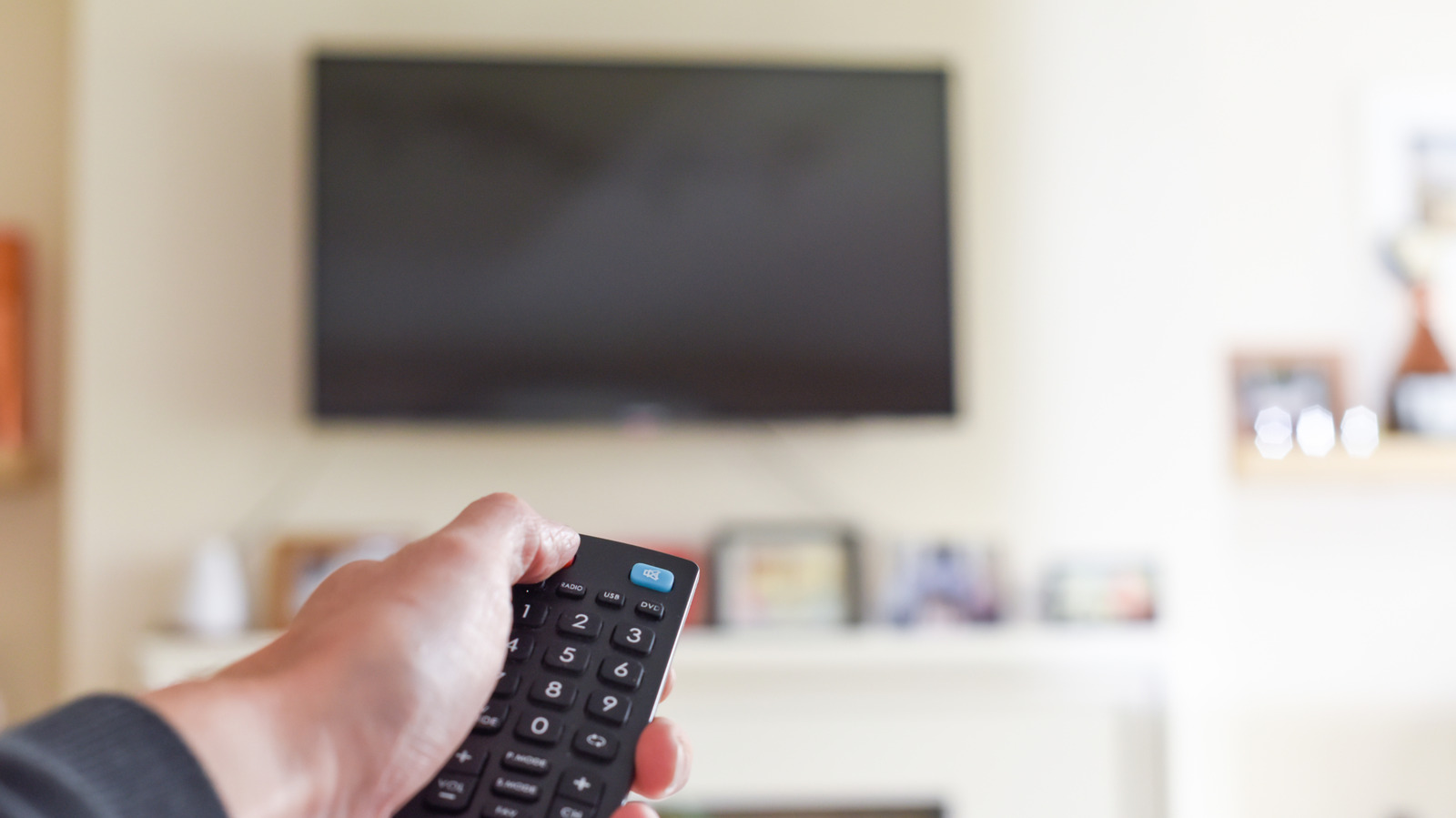 Hand holding a remote control toward a wall-mounted television in a softly blurred living room