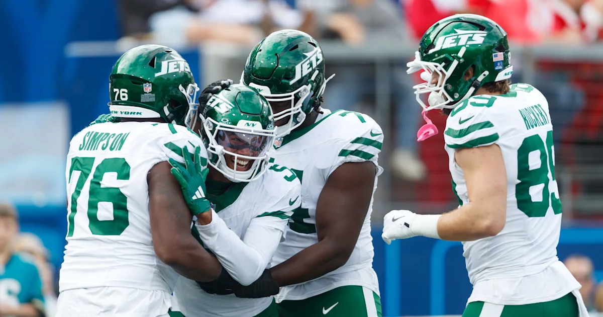 New York Jets players in white and green uniforms celebrate together, with a teammate standing nearby.