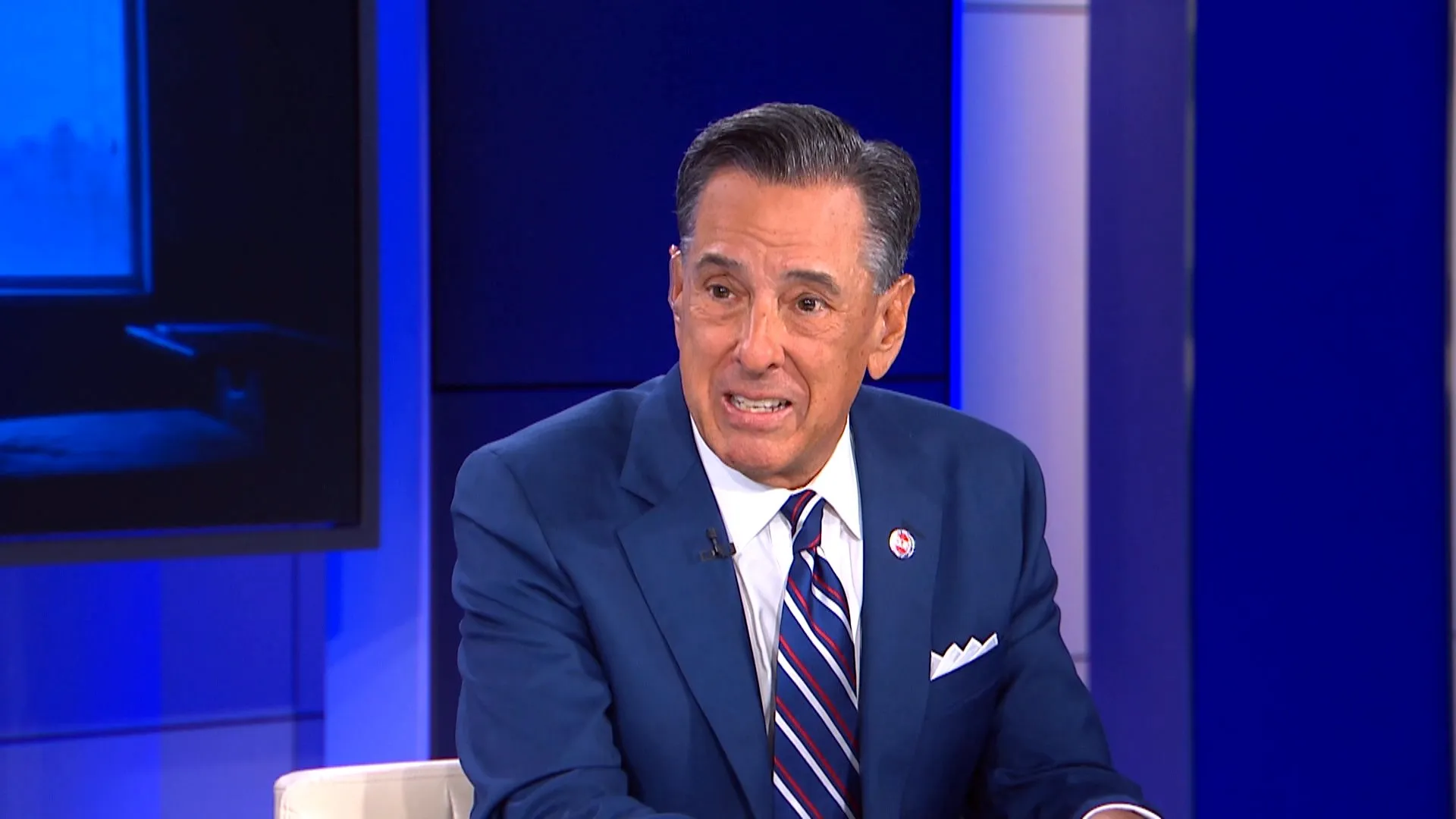 Man in a navy suit and striped tie speaking during a televised studio interview