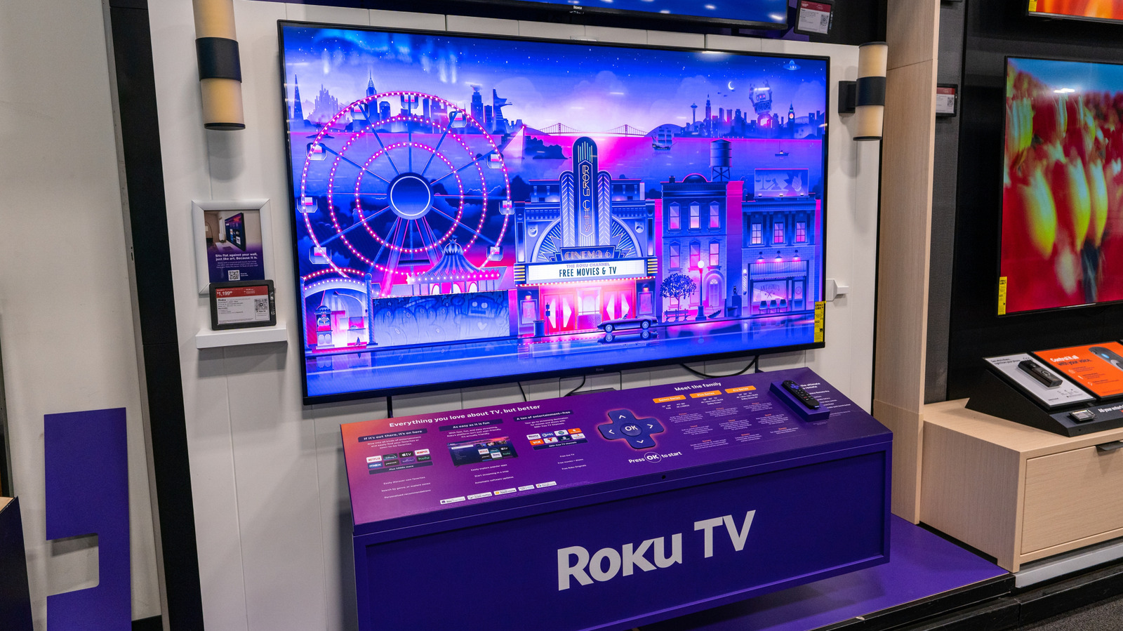 A Roku TV display in an electronics store shows colorful city artwork above a purple Roku-branded demo counter.