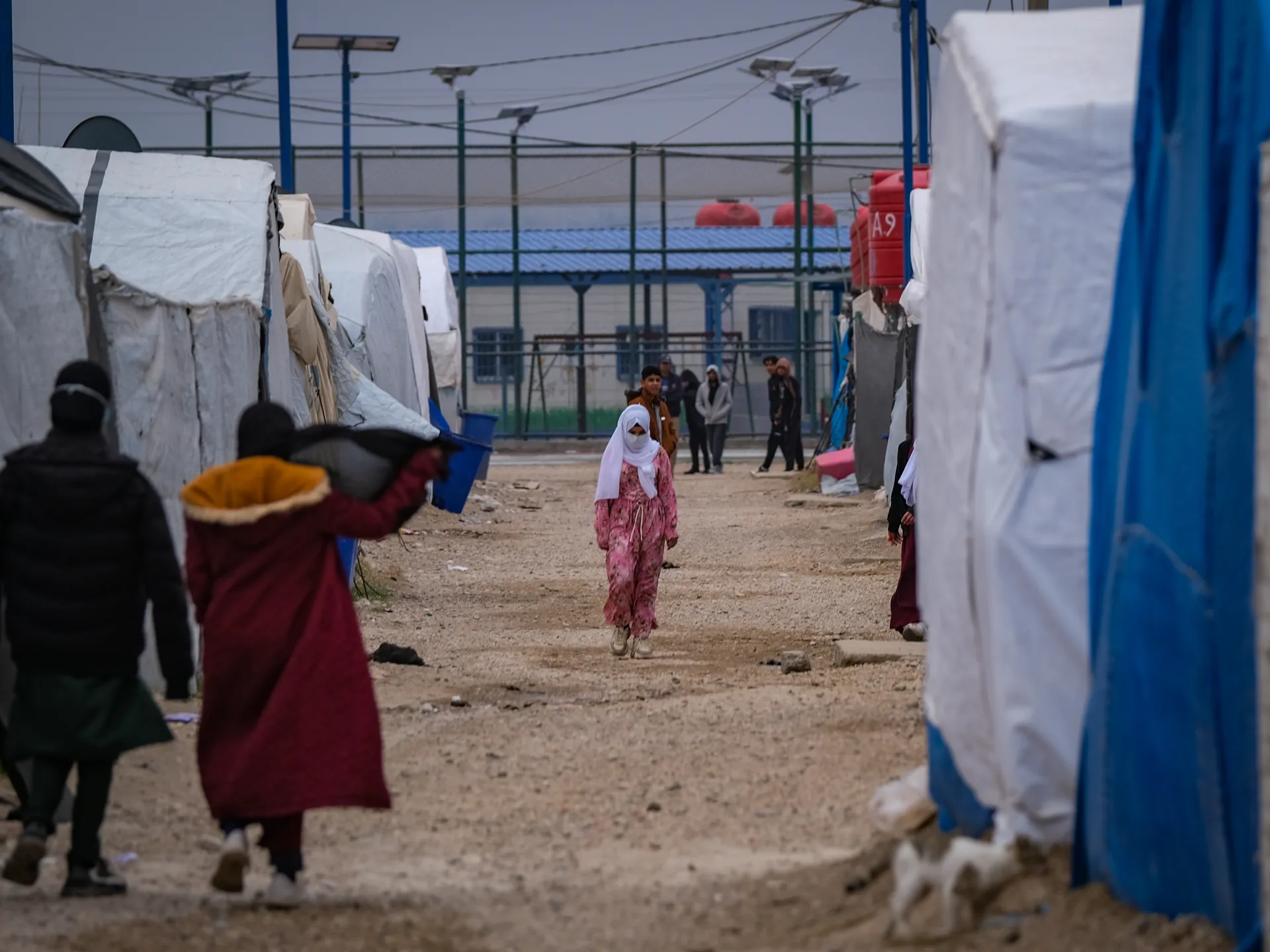 People walk between makeshift tents in a muddy camp, with solar streetlights overhead.