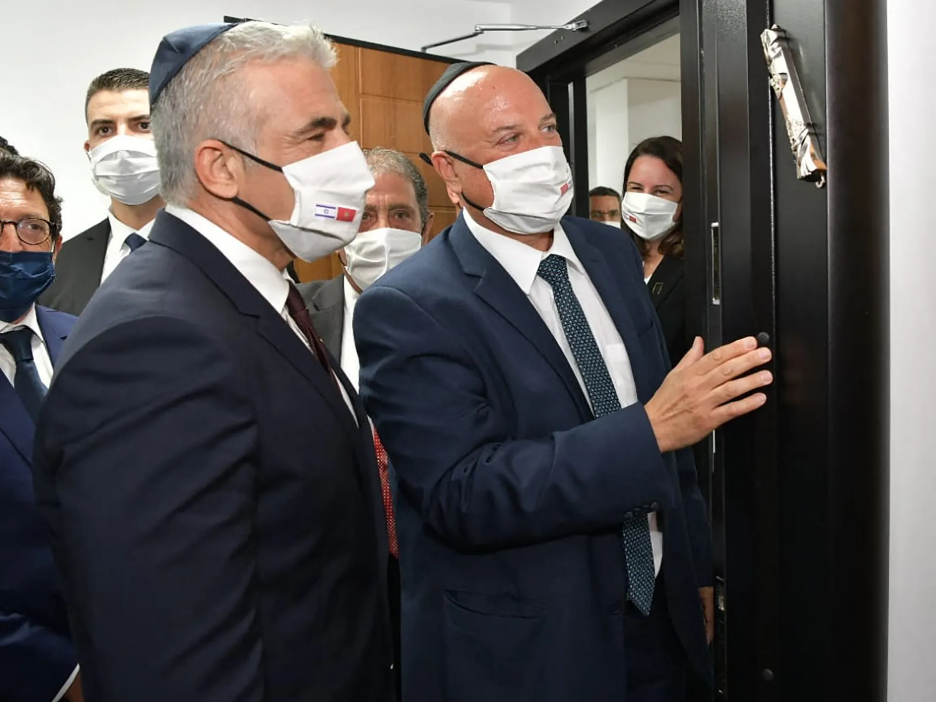 Masked officials in suits stand together near a doorway during a formal indoor gathering.