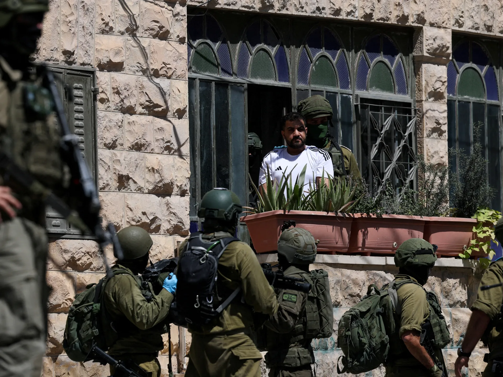 Armed soldiers escort a man from a stone building amid potted plants and barred windows