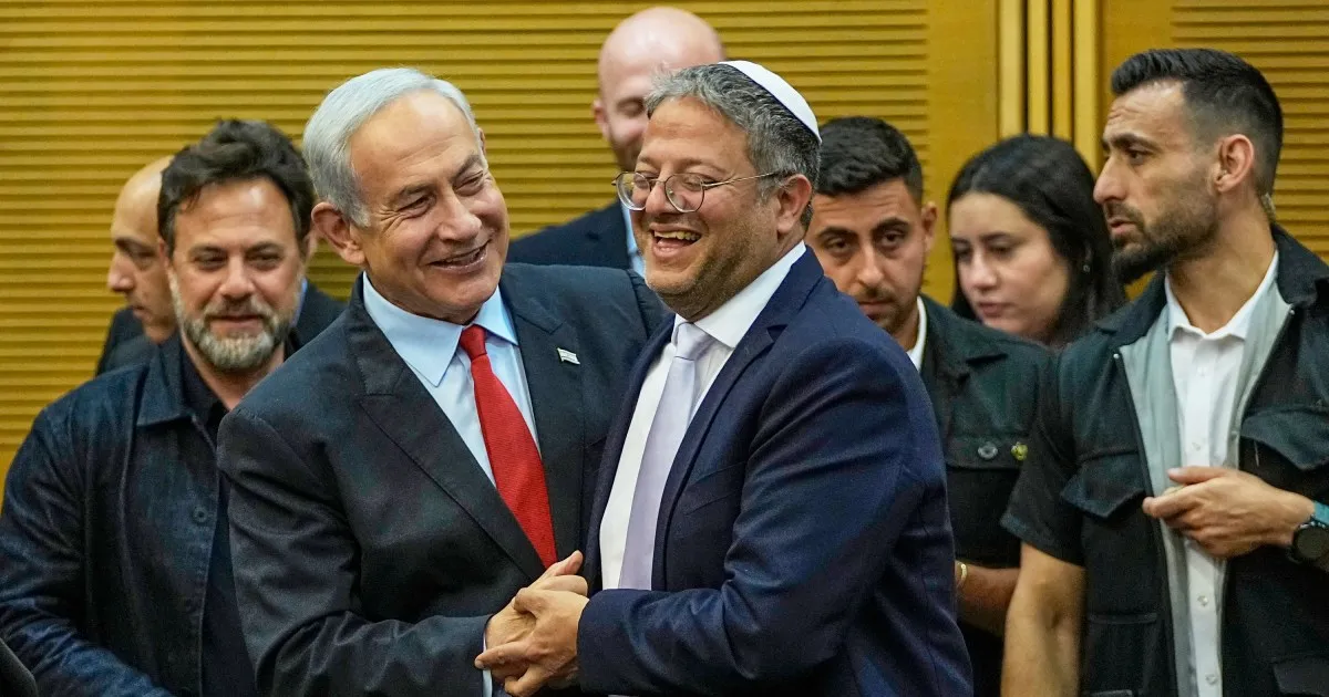 Two men in suits smile and clasp hands as several people stand behind them indoors.