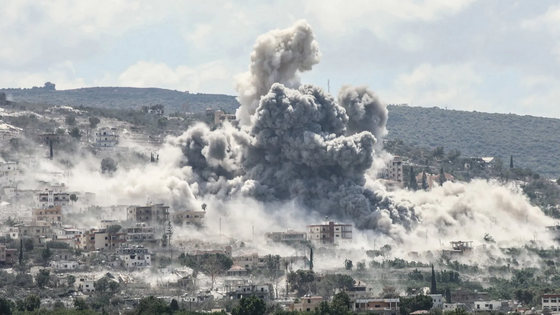 Large gray smoke plume rises over a densely built hillside town after an explosion.