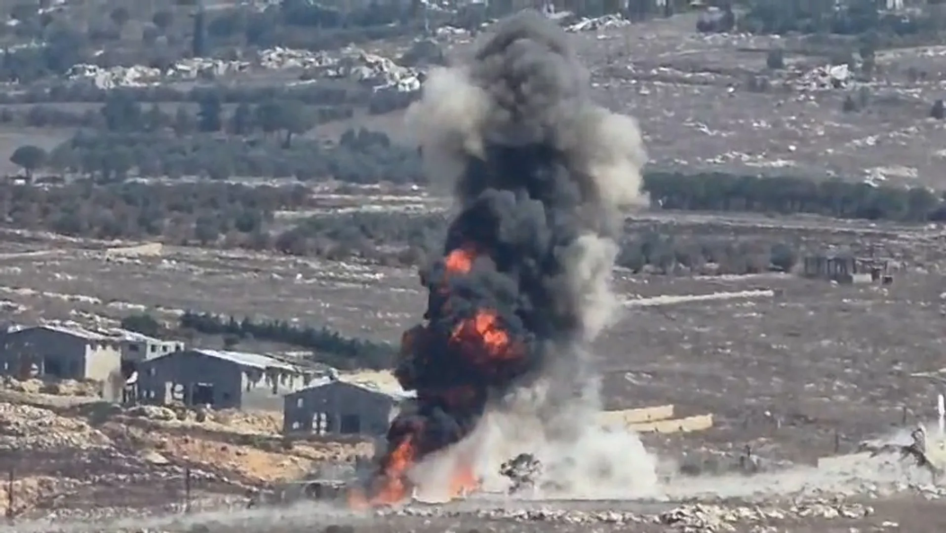 Large explosion with flames and thick smoke rising near damaged buildings in a barren landscape