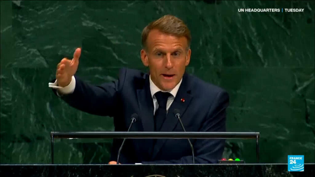 A suited man gestures behind a lectern against a dark green backdrop.