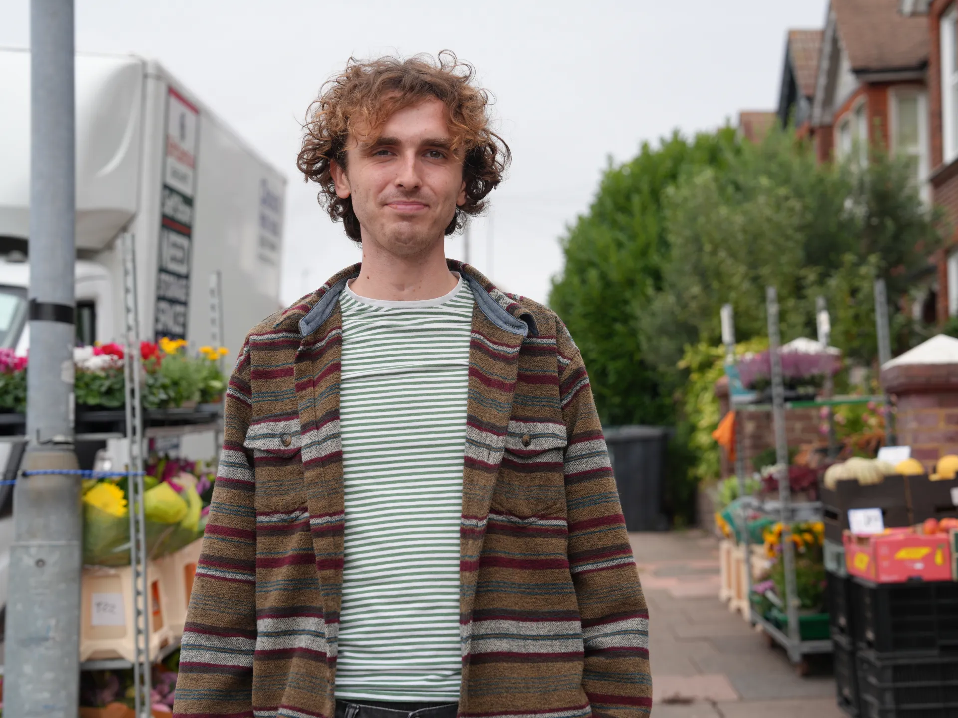Curly-haired man wearing a striped shirt and patterned jacket stands on a flower-market street.