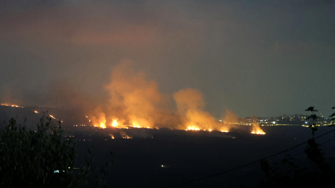 Nighttime fires burn along a dark hillside beneath thick smoke, with distant lights visible.