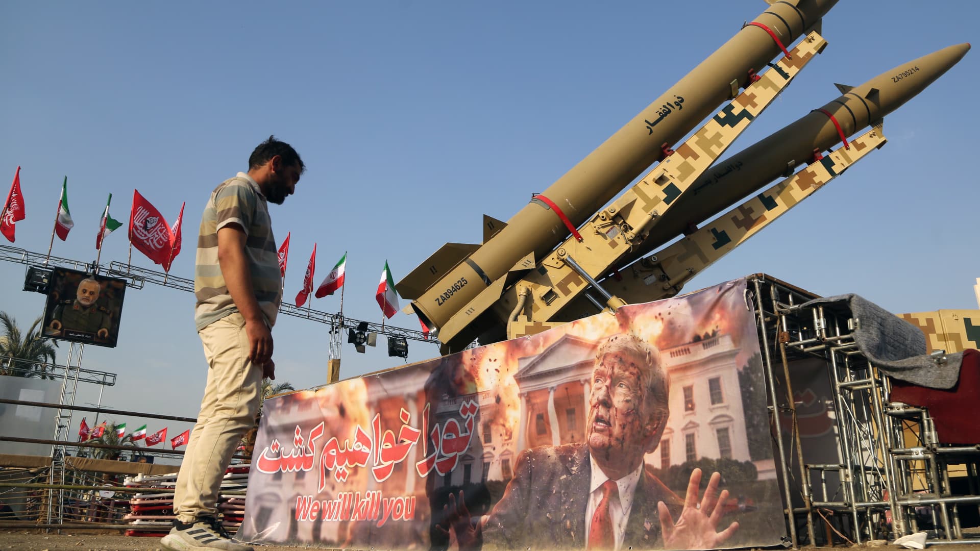 Man stands beside displayed missiles, flags, and a banner depicting Donald Trump at an outdoor gathering.