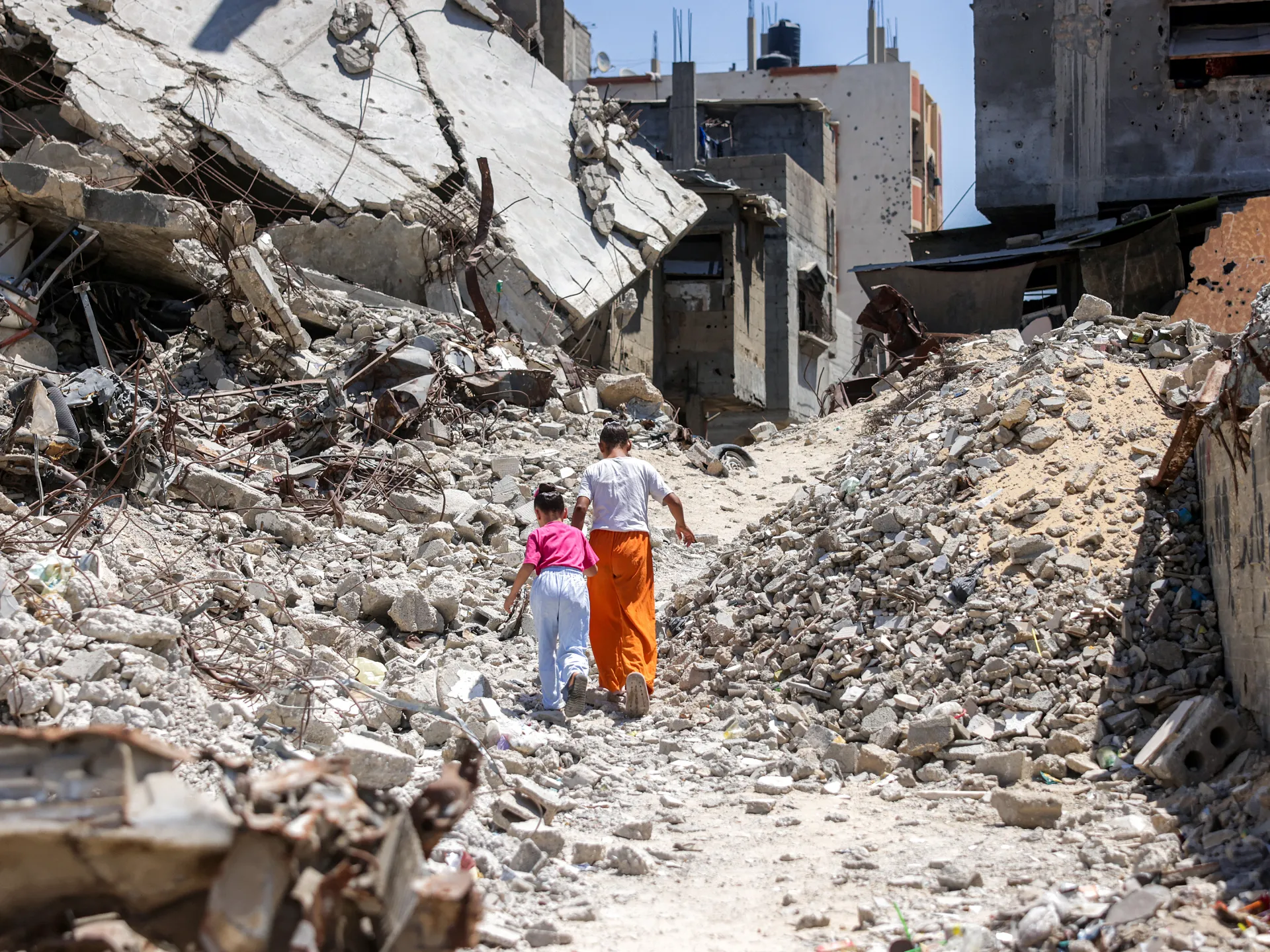 Two people walk through extensive rubble beside heavily damaged concrete buildings.