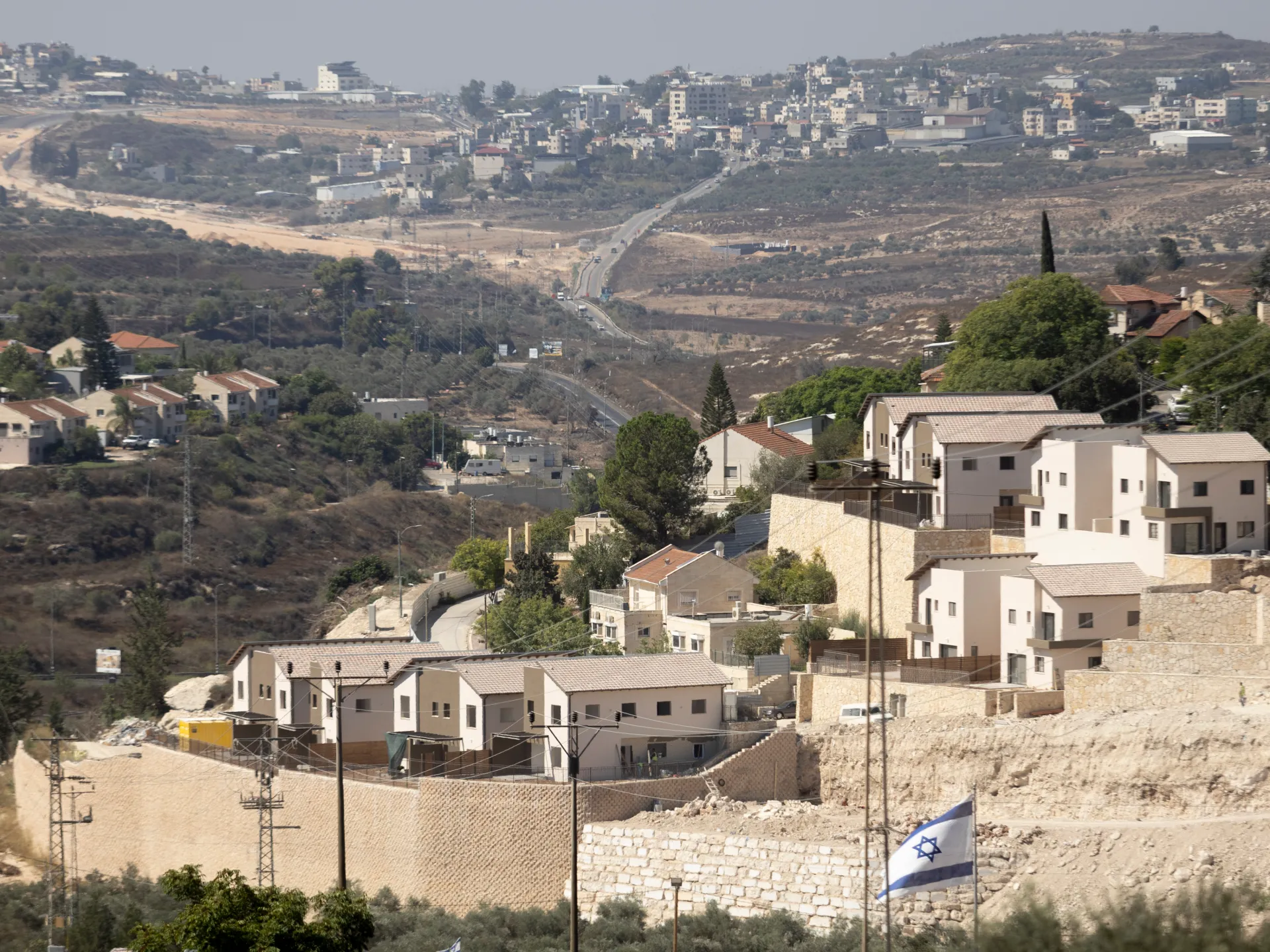 Israeli flag hangs beside stone walls and modern homes on a dry hillside.