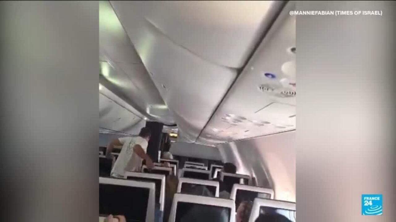 A passenger leans over a row of seats inside an airplane cabin with overhead bins open.