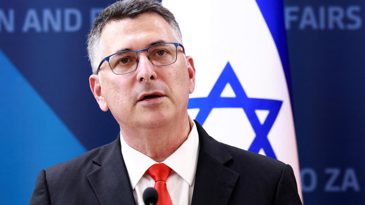 A bespectacled man in a dark suit and red tie speaks before an Israeli flag.