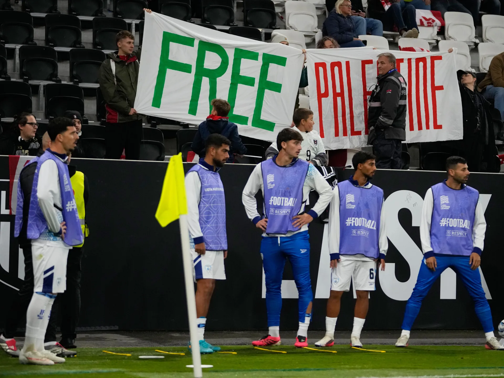 Football players stand pitchside before spectators holding a banner with large green and red lettering.