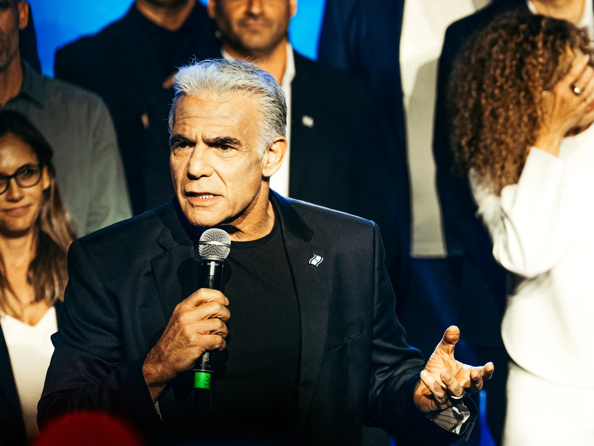 Silver-haired man in a dark blazer speaks into a microphone before a group of people.