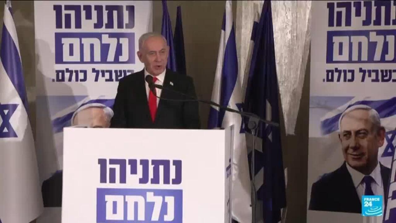 A suited man in a red tie speaks at a podium between Israeli flags and Hebrew campaign posters.