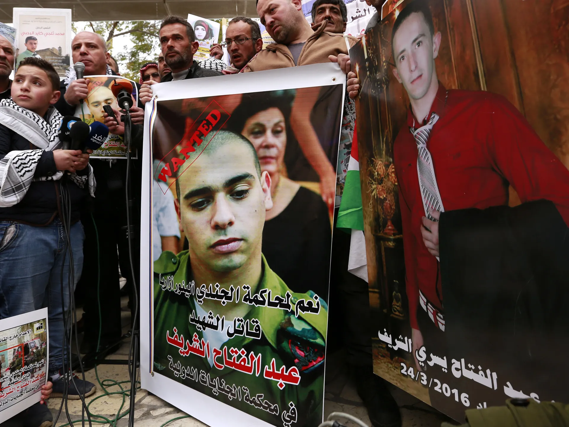 Protesters hold portraits and posters during a demonstration, with microphones visible in the foreground.