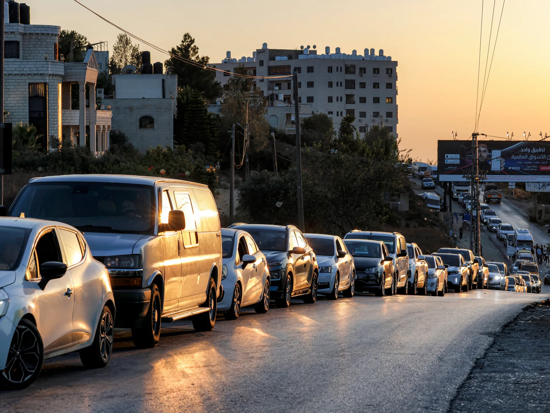 A long line of cars waits on a road beneath hillside buildings at sunset.