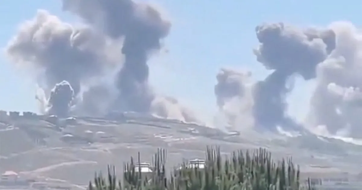 Large plumes of smoke rise above a barren ridge with tall vegetation in the foreground