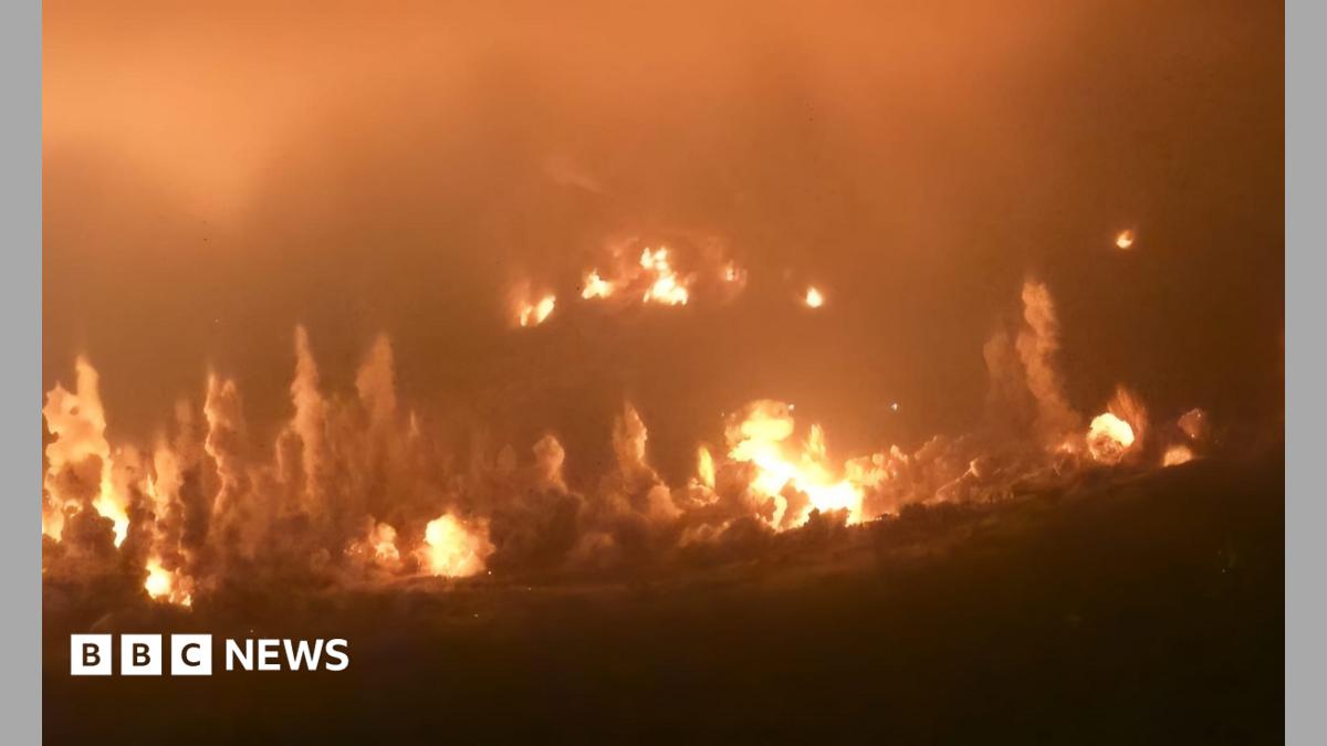 Nighttime fires and smoke rise across a dark landscape under an orange, hazy sky.