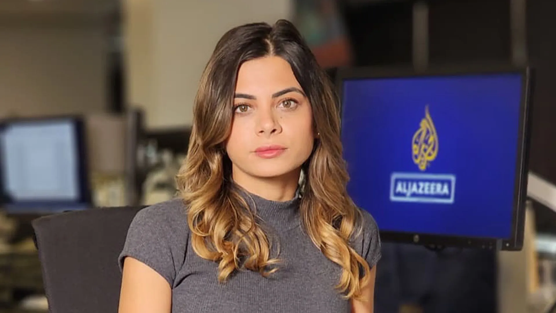 Woman with long brown hair seated indoors beside a monitor displaying the Al Jazeera logo