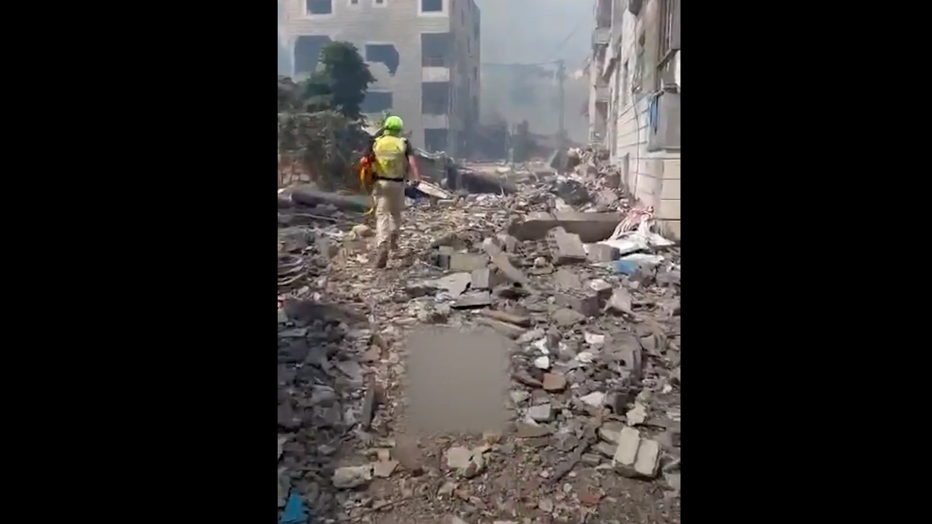 Emergency responder walks through rubble between damaged buildings amid smoke and destruction.