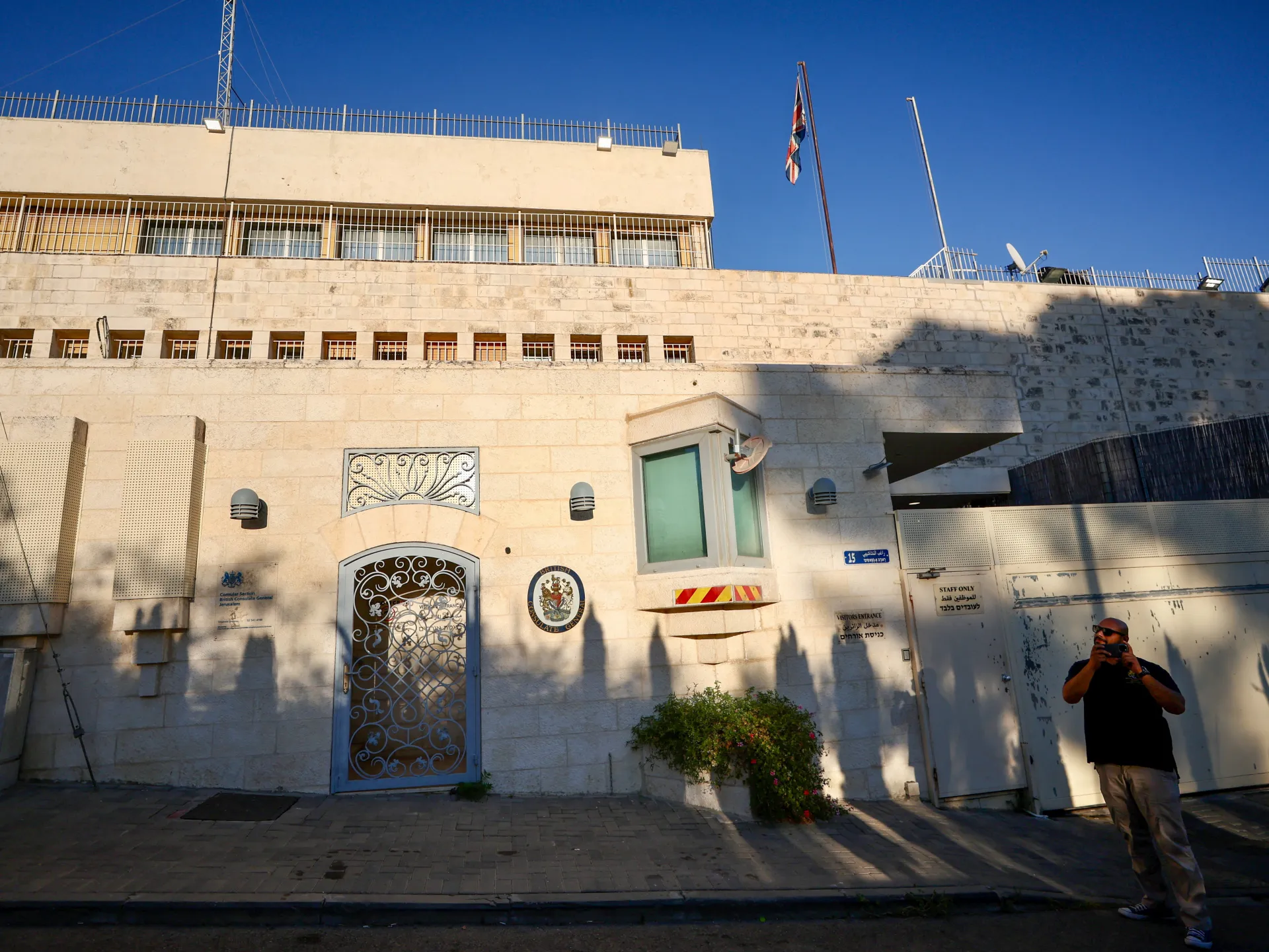 Exterior of British Consulate General Jerusalem with barred windows, flags, entrance gates, and a visitor outside.