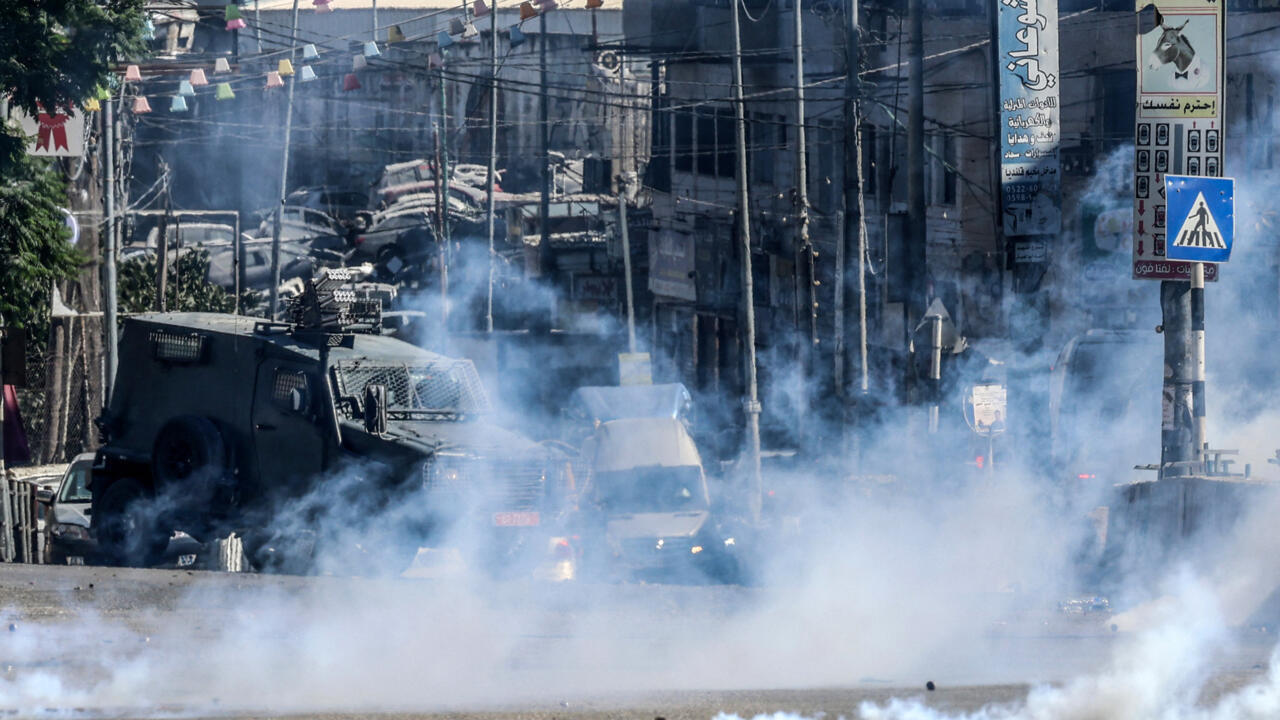 Armored vehicle moves through heavy smoke on a dense urban street lined with utility wires.