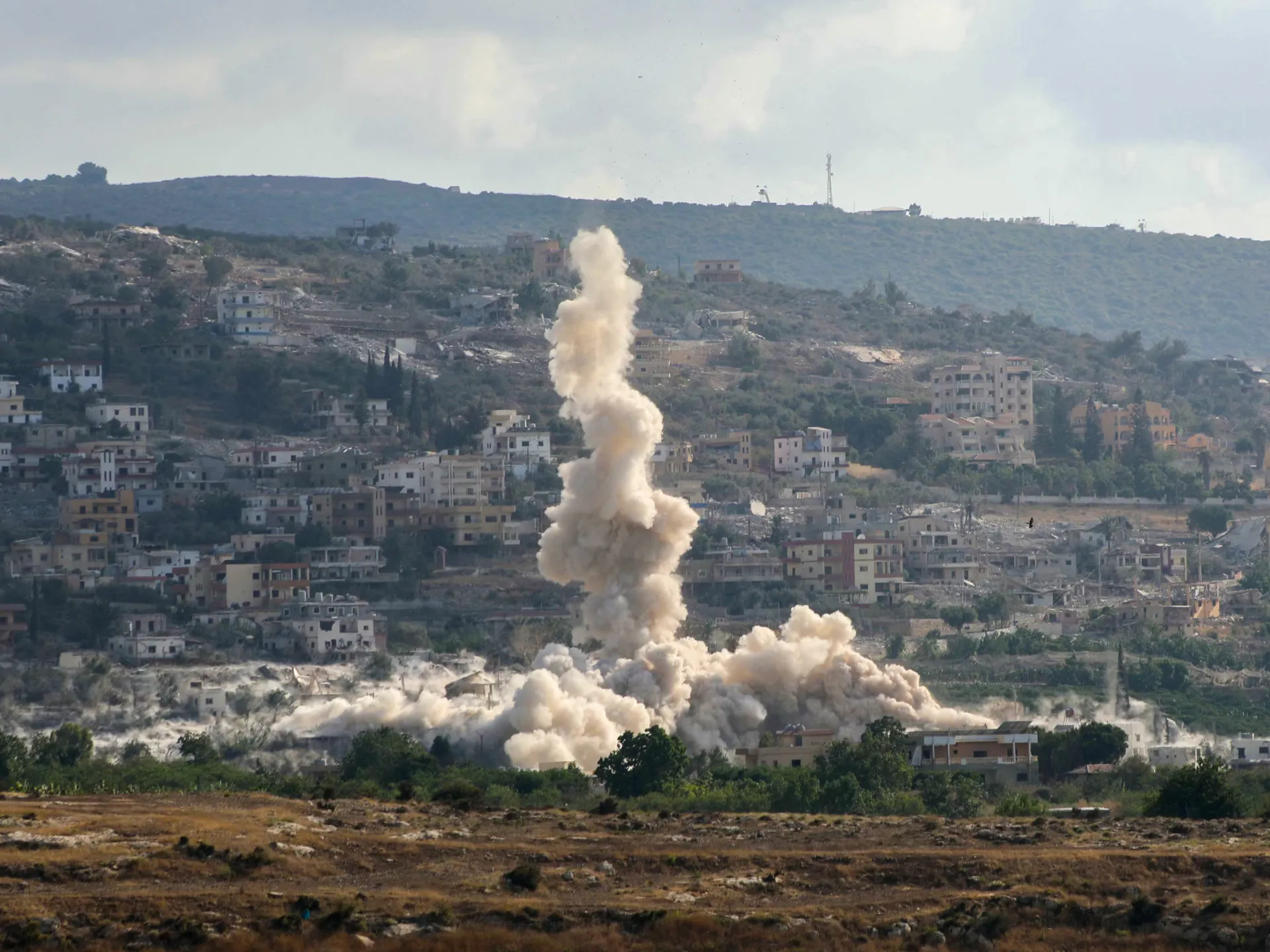 A large explosion sends a column of dust above a densely populated hillside settlement.