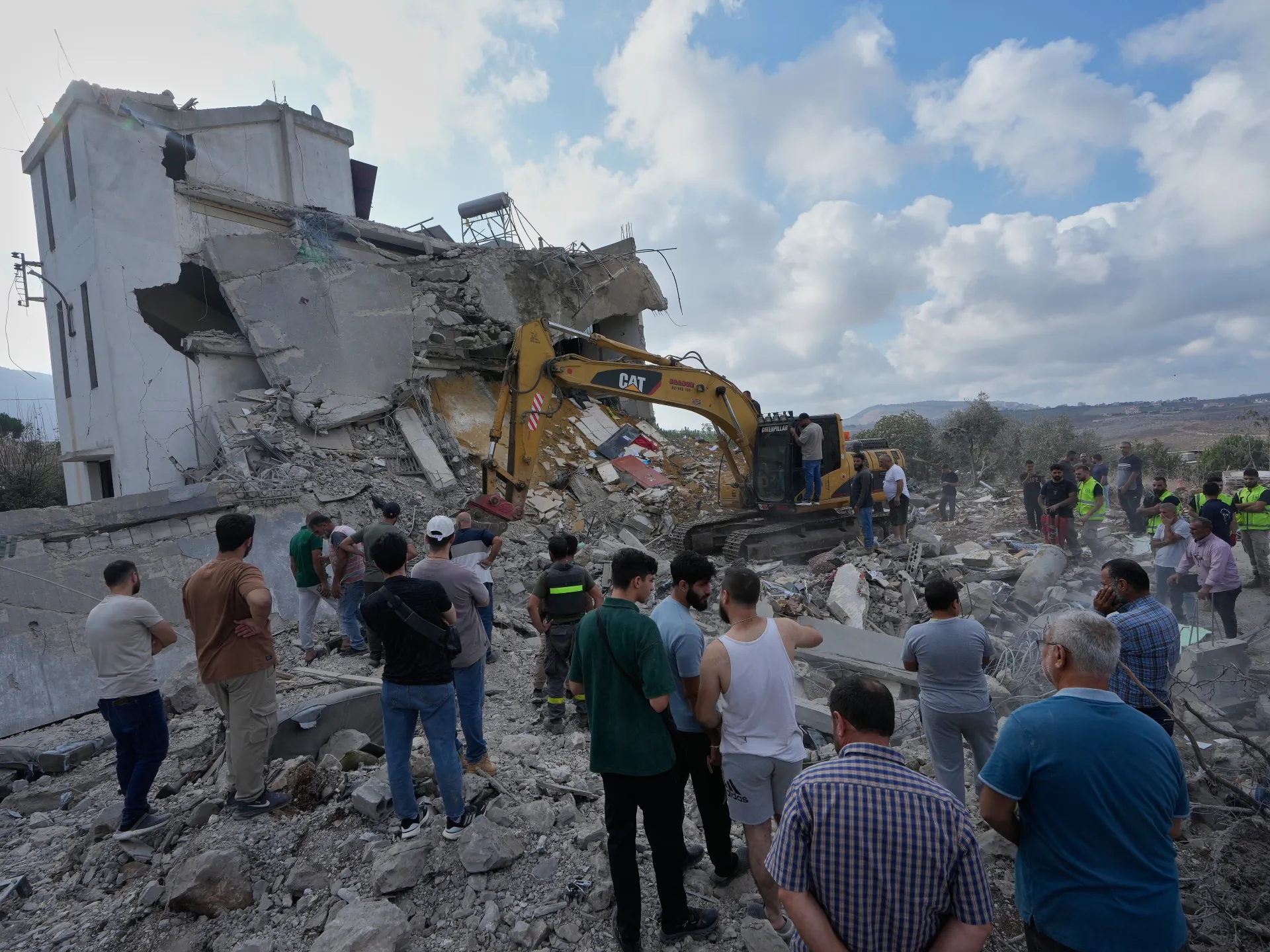 Crowd gathers beside an excavator clearing rubble from a heavily damaged building.