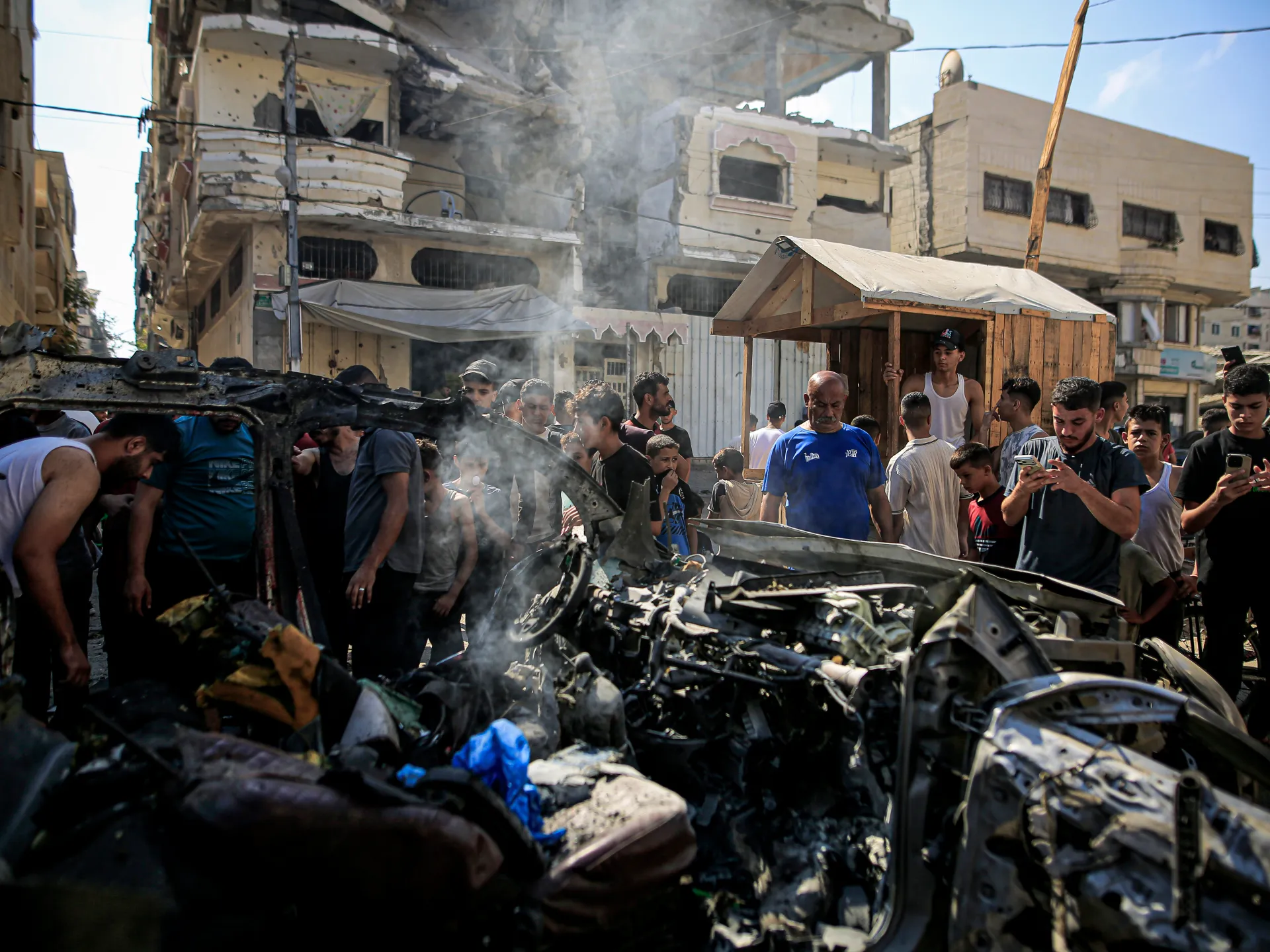 Crowd gathers around a smoldering wrecked vehicle amid heavily damaged buildings
